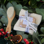 Cosy Cottage baby products including a wooden brush, washcloth, and soap on a green leafy background.