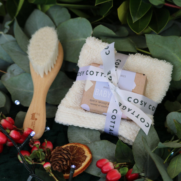 Cosy Cottage baby products including a wooden brush, washcloth, and soap on a green leafy background.