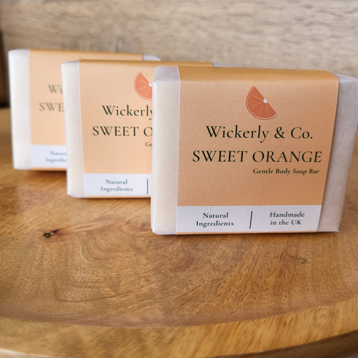 Three wrapped white label soap bars made by Cosy Cottage for 'Wickerly & Co. Sweet Orange' arranged on a wooden surface, with the front bar in focus and the others softly blurred in the background.