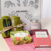 geranium and bergamot soap, lavender hand and body cream, rhubarb milk chocolate and green woollen socks positioned in front of a Cosy Cottage gift box