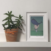 framed cooperillo picture of a man with whale haircut standing in the sea, on a shelf next to a pot plant