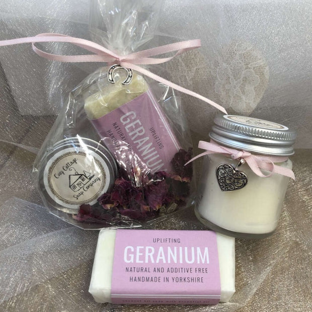 geranium travel soap and soy wax candle in front of bundle of soap and lip balm in biodegradable packaging with a pink ribbon 