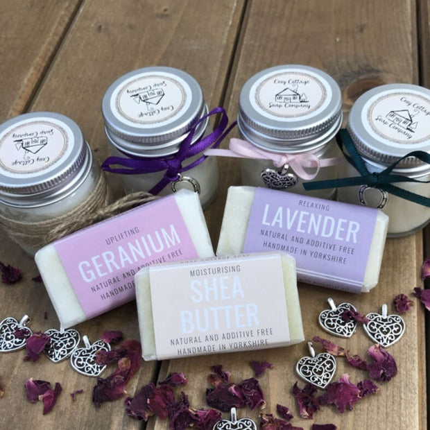 fragranced travel soaps and soy candles arranged on a wooden bench top with rose petals